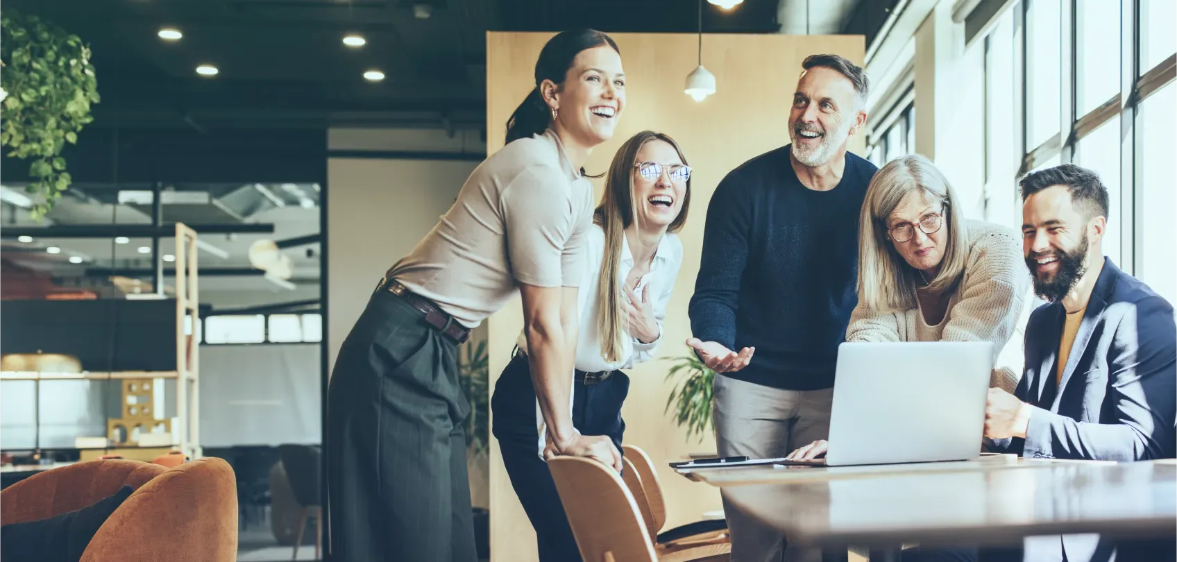 Equipo de trabajo diverso reunido alrededor de una laptop, sonriendo y colaborando en un espacio de oficina moderno.