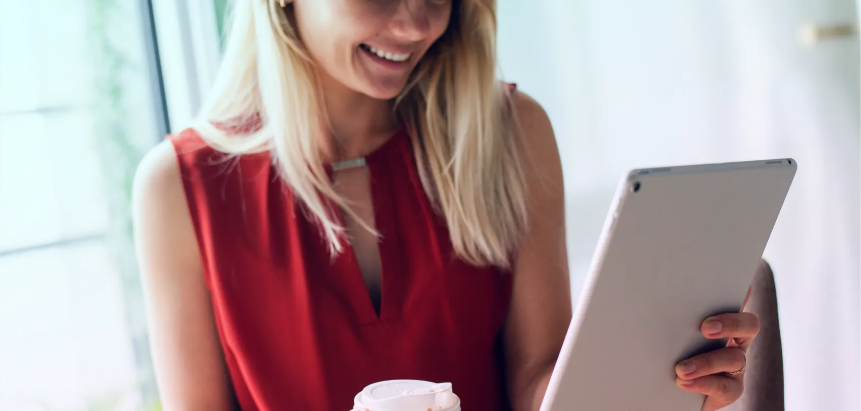 Mujer sonriendo mientras lee en una tablet y sostiene un vaso de café, en un entorno luminoso.