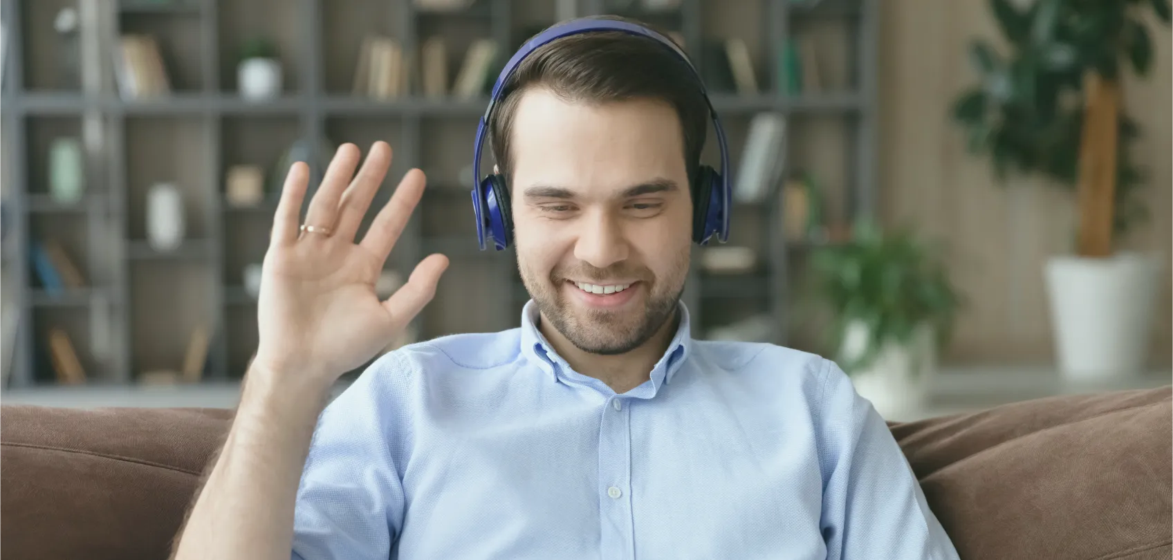 Hombre joven con auriculares azules saludando en una videollamada desde casa, con librero y plantas de fondo.